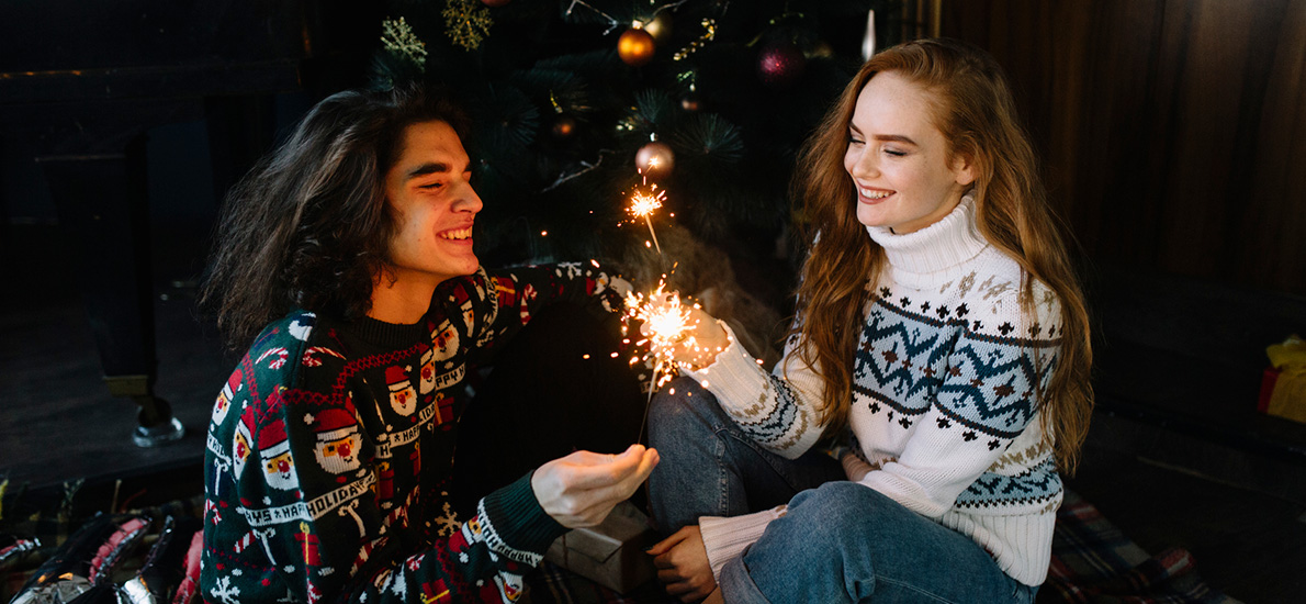 Couple in sweaters sitting in front of Christmas tree laughing holding sparkler