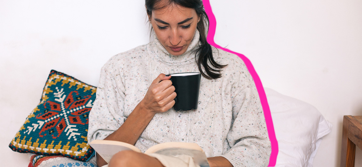 woman wearing a sweater reading a book and holding a mug