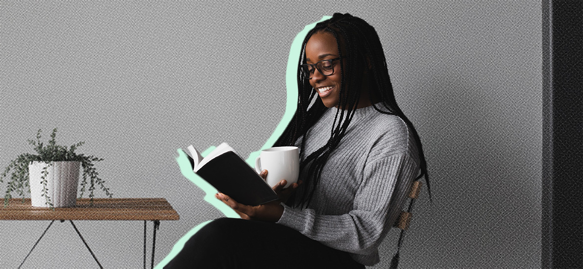 young Balck woman wearing glasses and a gray sweater, reading a book and holding a mug of coffee, smiling