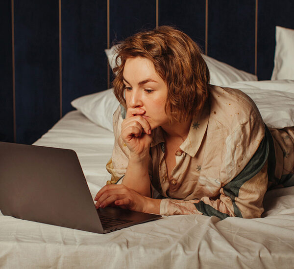 woman in pajamas laying on her bed has her hand in her face, in thought