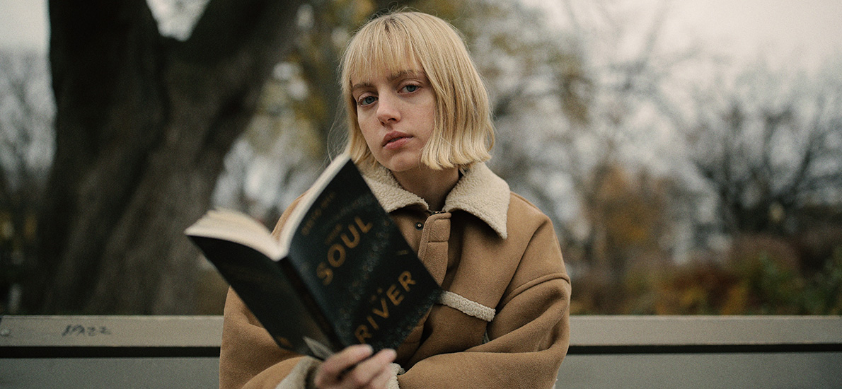 girl with blond bob and blunt bangs sitting outside on a bench, holding an open book