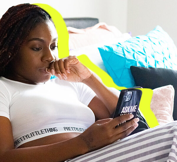 woman in a white PrettyLittleThing crop top and white and gray striped pants, sitting on her bed looking at her phone with her hand on her mouth