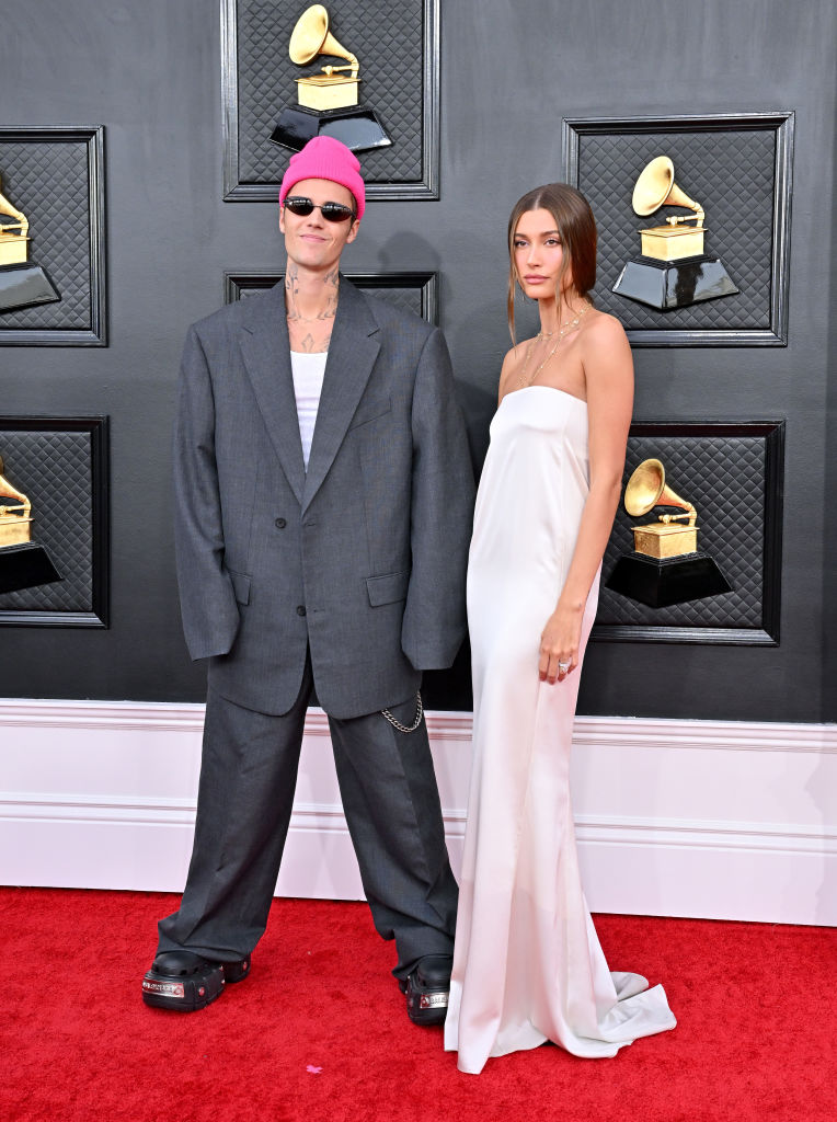 Justin and Hailey Bieber on the 64th annual Grammys red carpet. Justin is wearing an oversized suit and neon pink beanie and Hailey is in a flowy white strapless gown.