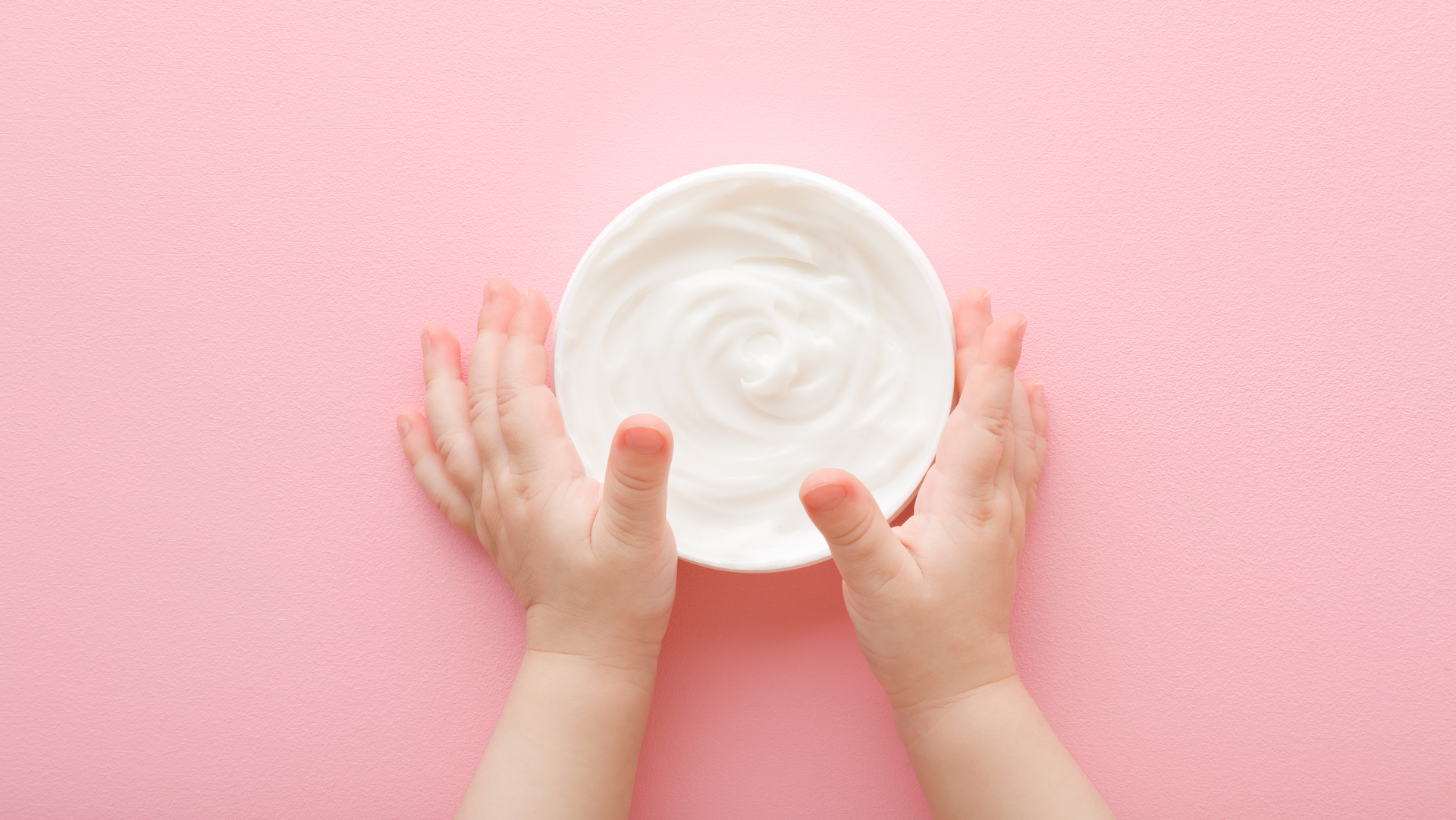 Baby girl little hands touching opened white cream jar on light pink table background.