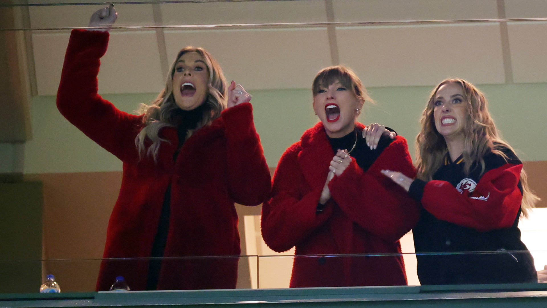 Taylor Swift in a red peacoat cheering with friends at a football game.