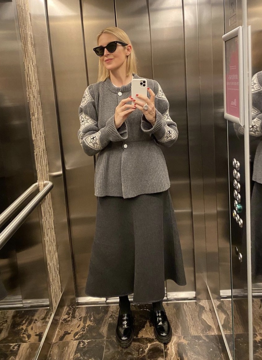 Kelly Rutherford taking a mirror selfie in an elevator mirror wearing a grey sweater and skirt