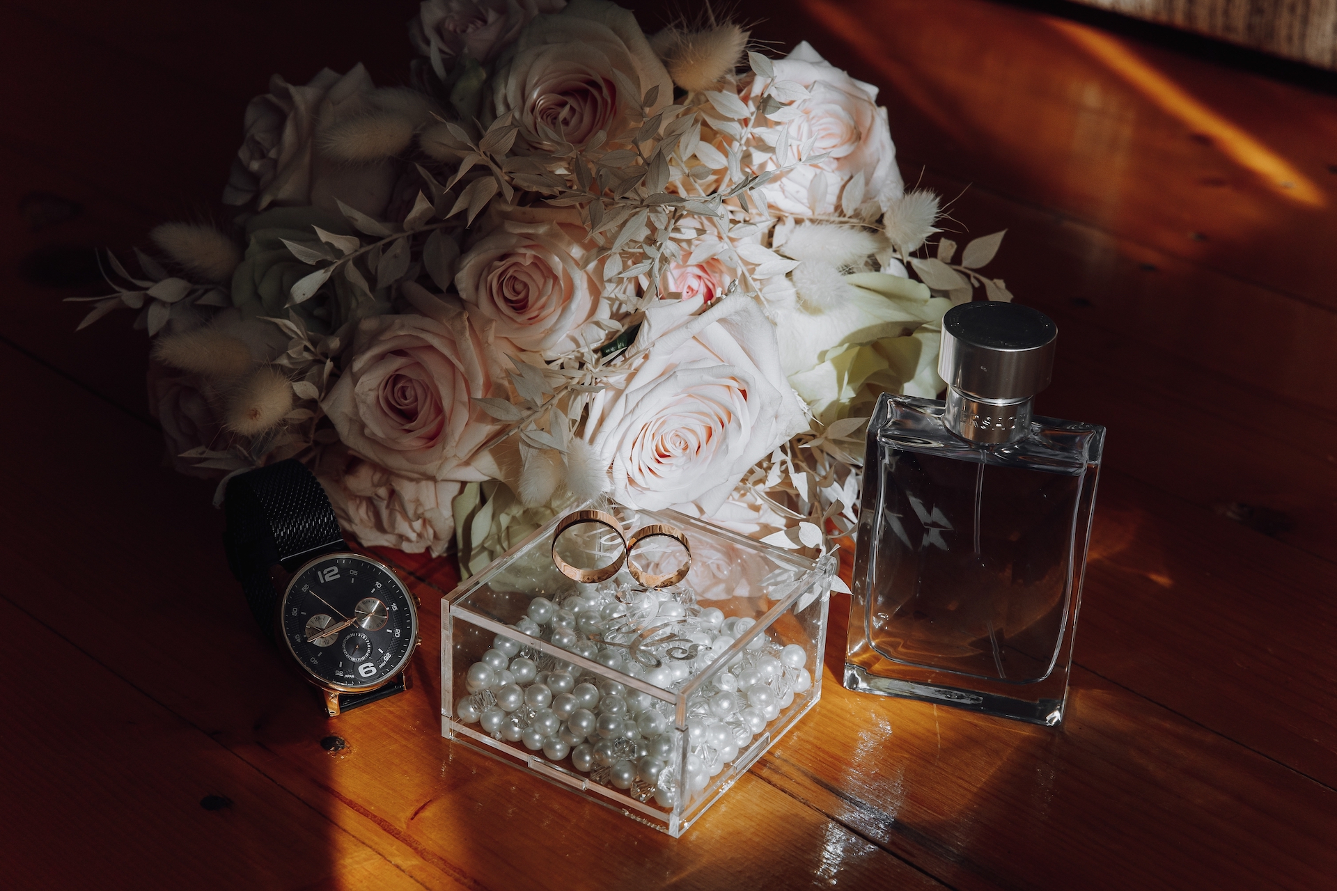 A bouquet of flowers and a watch are displayed on a wooden table. The watch is a silver one with a black band. The flowers are pink and white and arranged in a vase. Scene is elegant and sophisticated